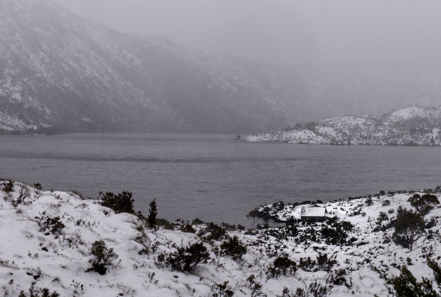 Snow-covered shoreline with dark mountain lake and misty mountain backdrop in winter landscape.