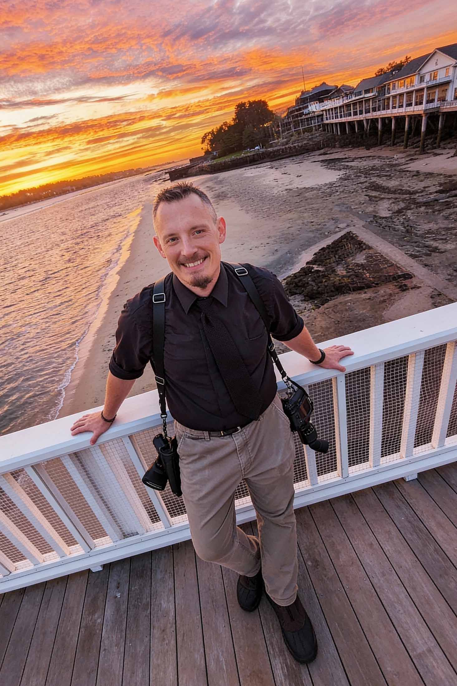 Candid headshot of wedding photographer Gregory Hitchcock, relaxing on a dock over looking the ocean with the sun setting beyond.