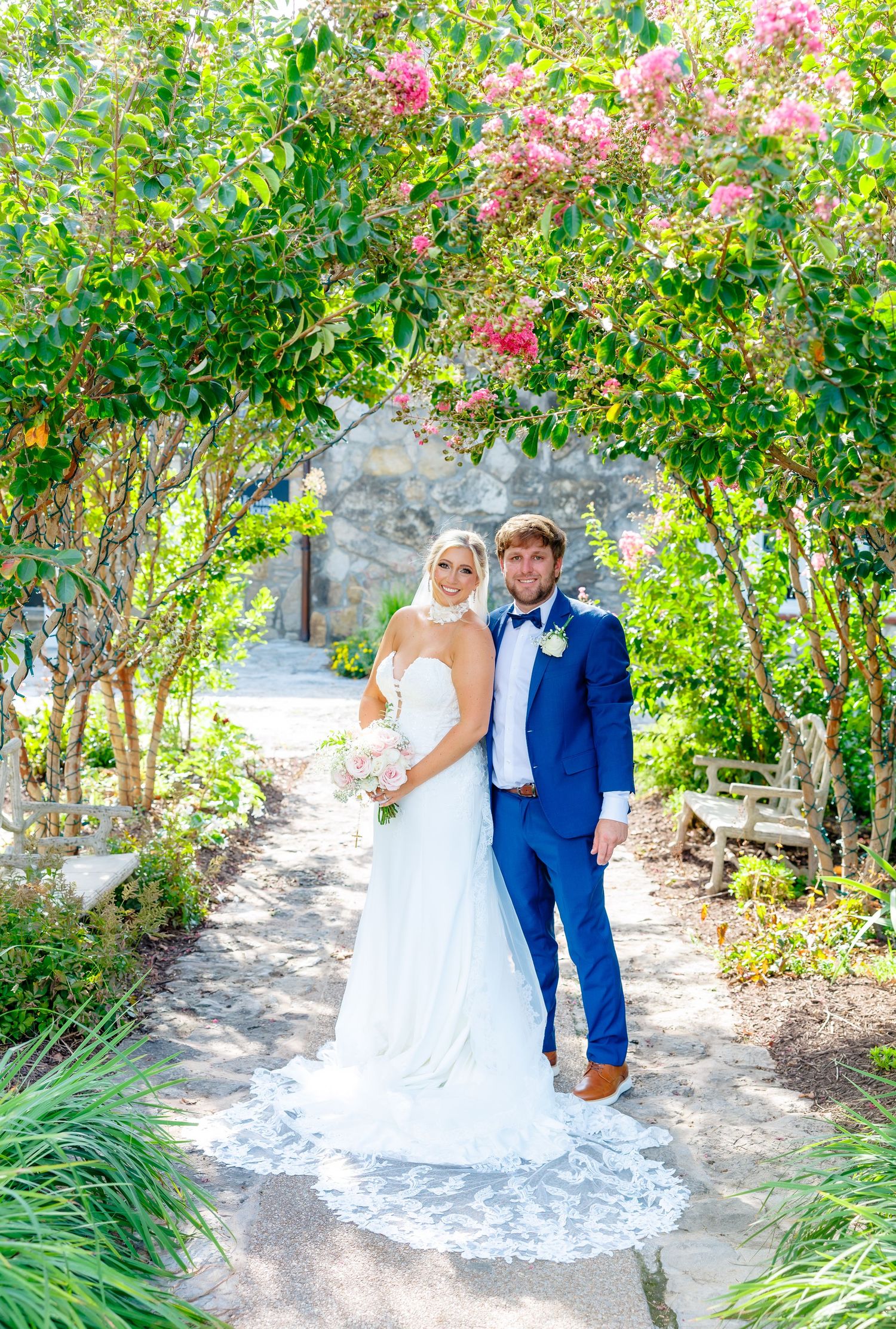 Bride and groom pose under flowering archway in lush garden, bride in white gown, groom in blue suit.