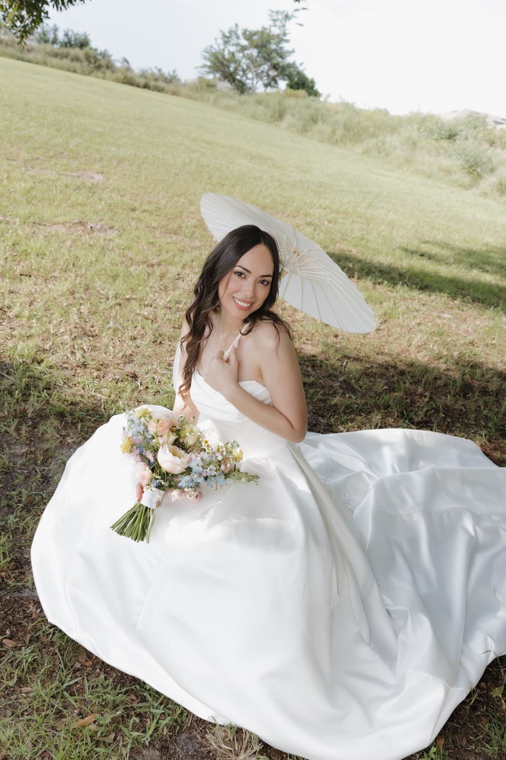 Smiling bride in white gown sits on grass holding bouquet and parasol umbrella outdoors.