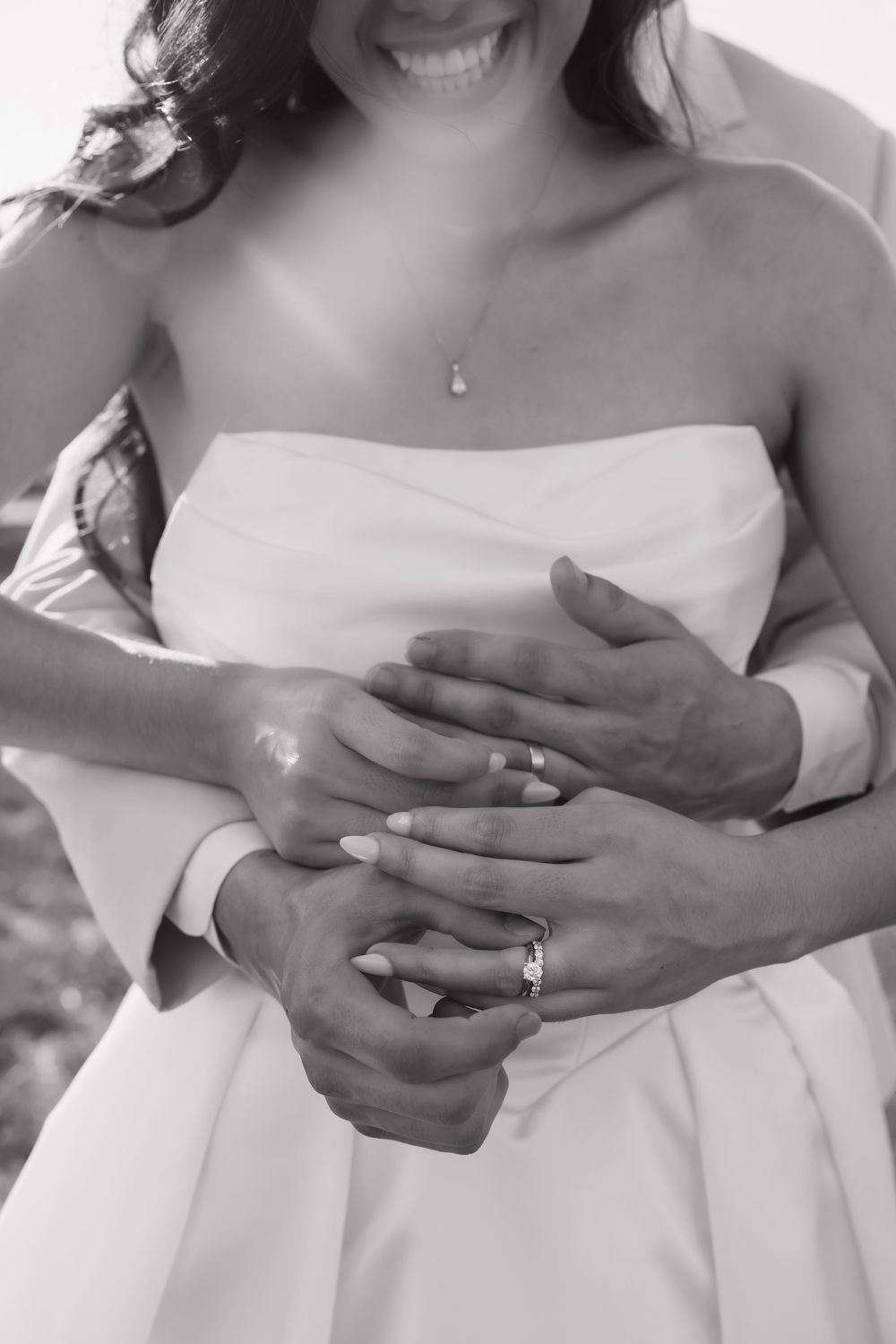 Black and white photo of bride and groom holding hands, showing wedding ring, bride in strapless gown with pearl necklace.