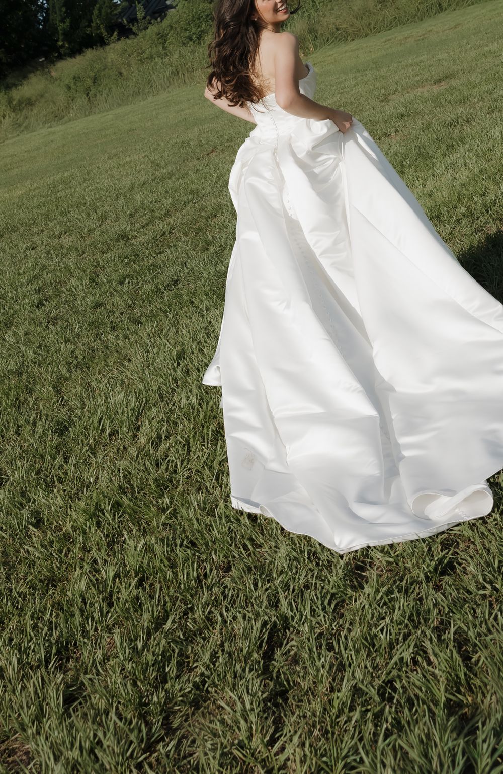 Bride in a flowing white strapless satin wedding gown walking through a lush green field, holding up her dress train.