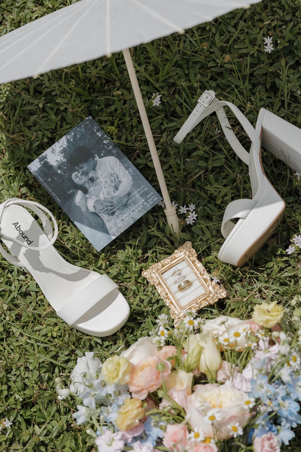 Bridal flat lay with white heels, bouquet, rings in gold frame, parasol on green grass.
