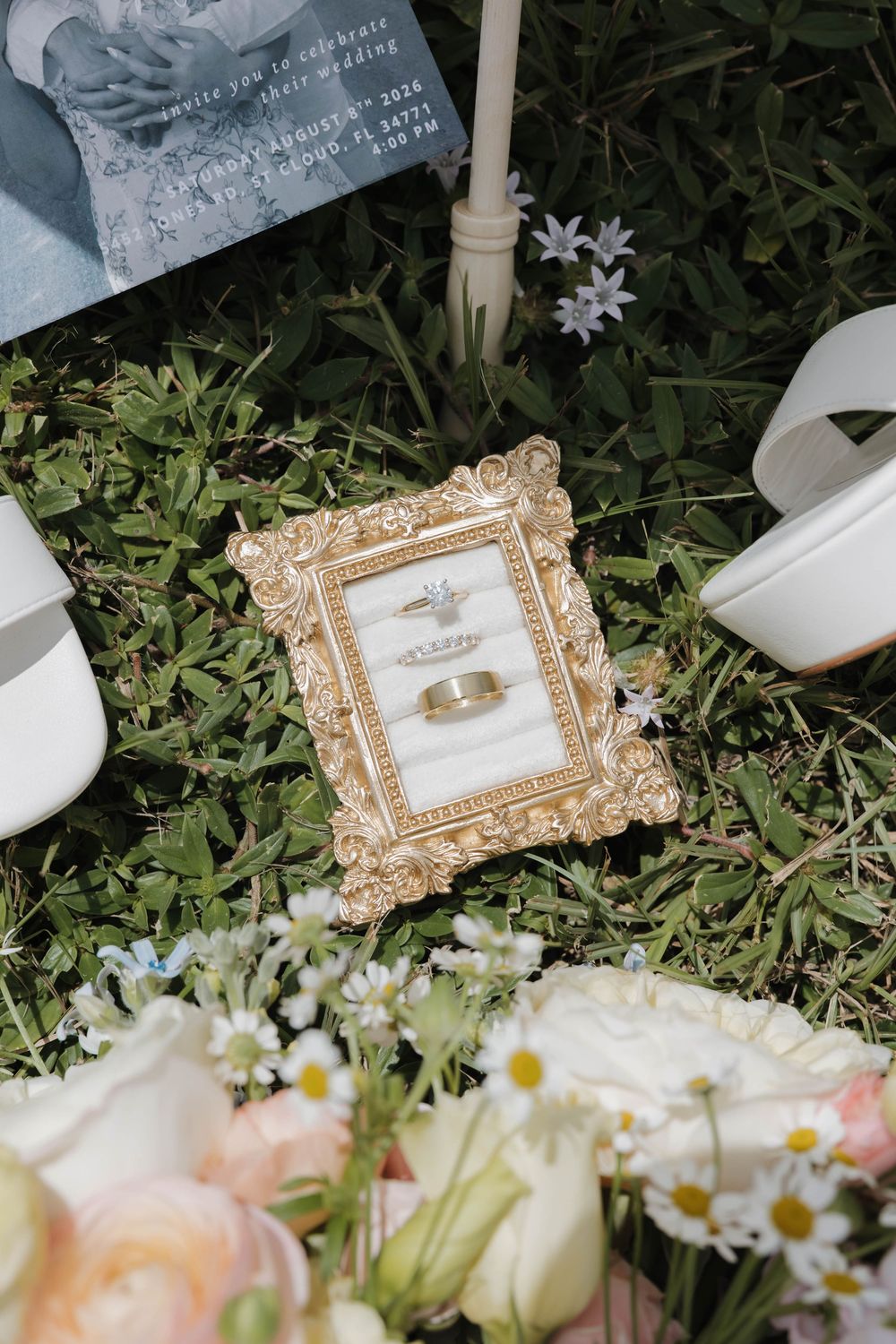 Gold ornate frame displaying wedding rings on grass, surrounded by bridal bouquet, white heels, and wedding invitation.