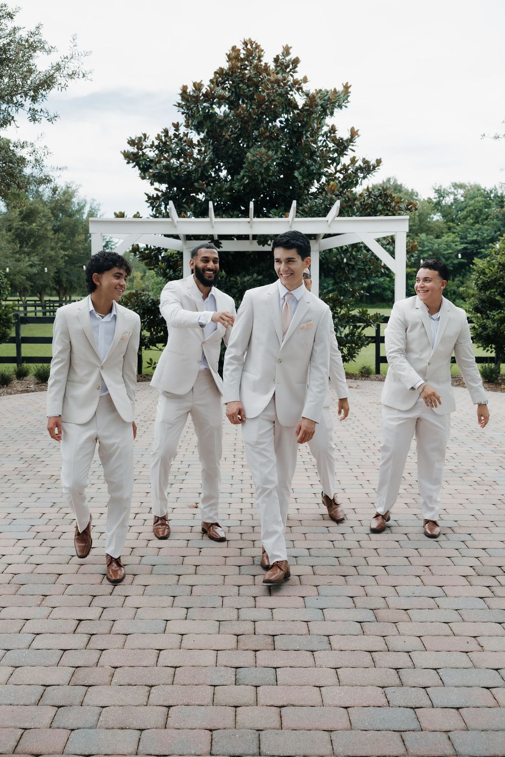 Four groomsmen in matching cream suits walk confidently on a brick patio at an outdoor wedding venue.