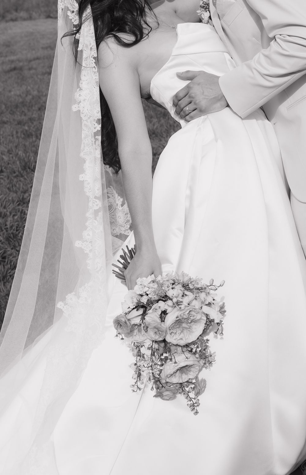 Black and white wedding photo of bride in strapless gown with lace veil holding floral bouquet, embraced by groom.