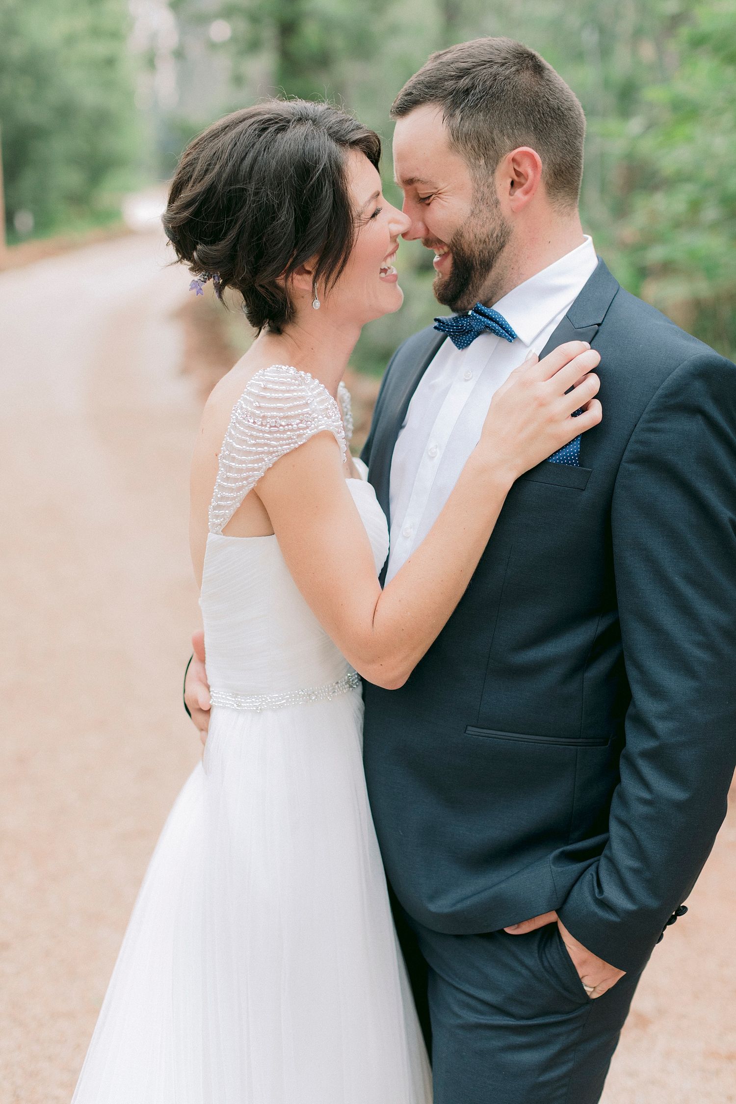 A bride in a white gown and groom in a navy suit with blue bow tie share a romantic moment on a tree-lined path.