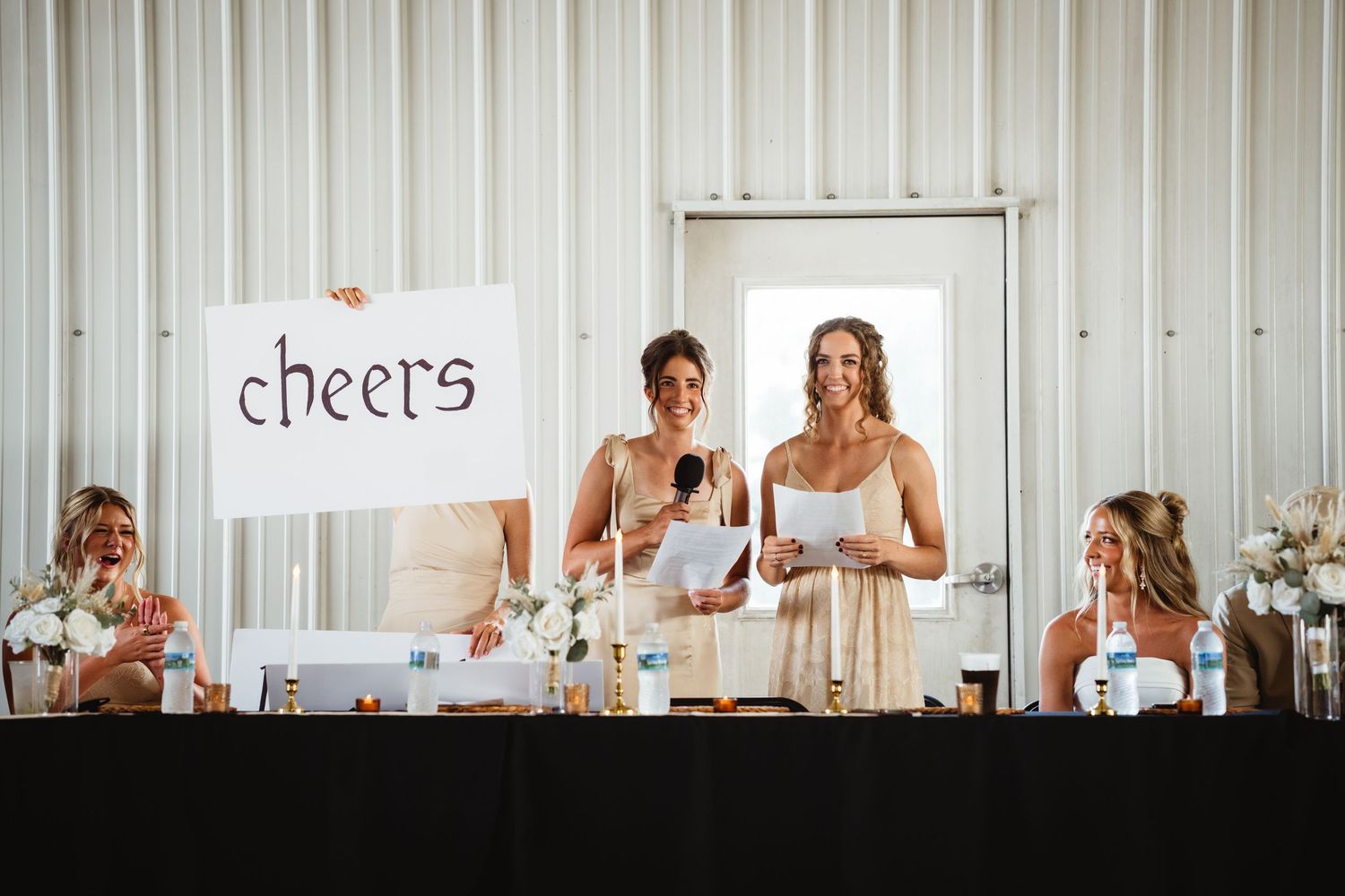 Bridesmaids giving a toast at a wedding reception, one holding a cheerful 'cheers' sign at the head table.