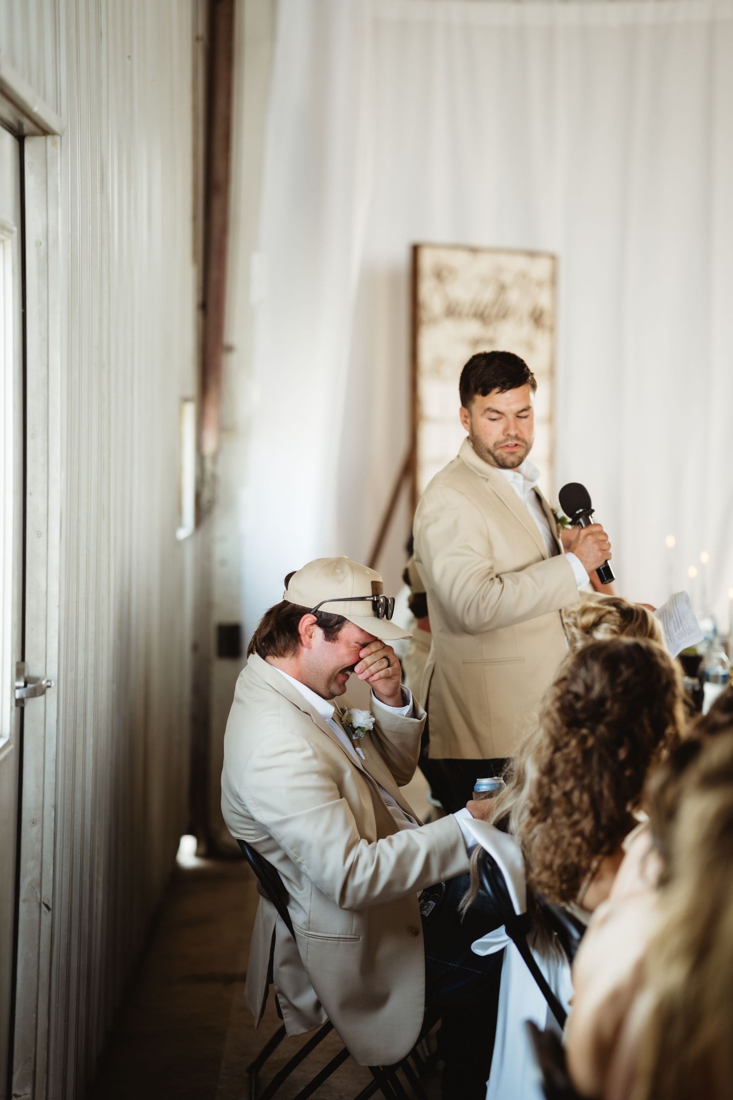 Groom laughing during best man wedding speech at elegant indoor reception venue.