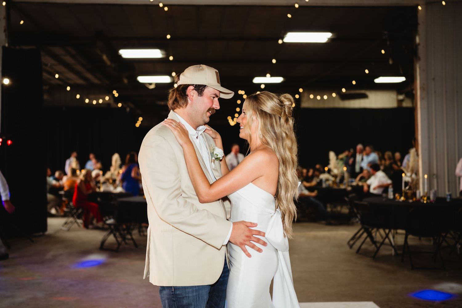 Bride and groom share first dance in rustic wedding venue with string lights and guests seated at tables in background.