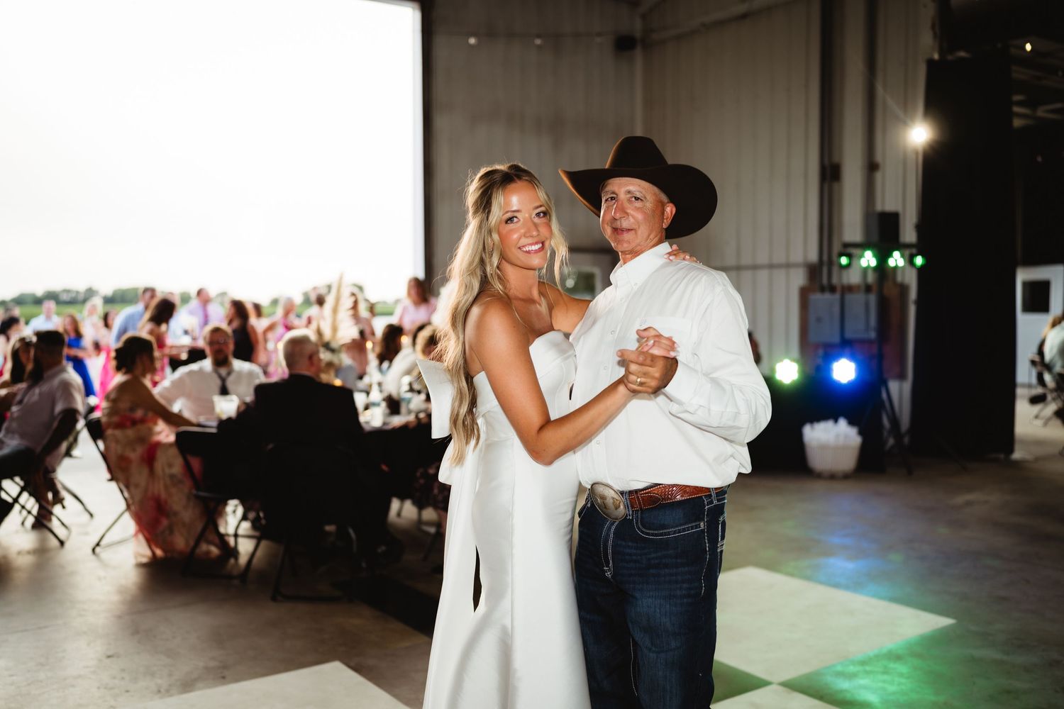 Bride and groom share first dance in rustic barn wedding venue with guests seated at tables in background.