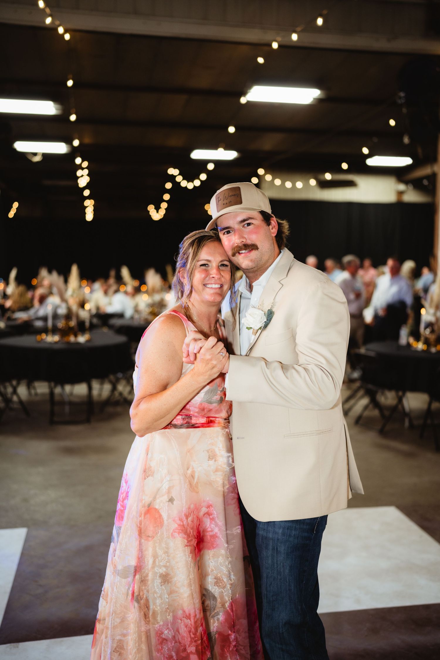 A couple poses together at a wedding reception venue decorated with string lights, the man wearing a cream blazer and trucker hat.