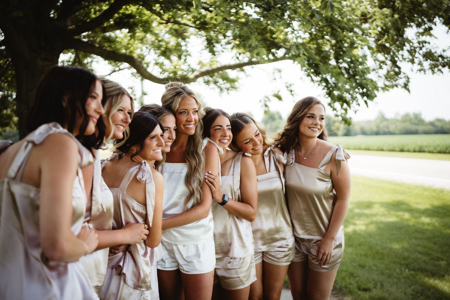 Seven women in matching white and beige outfits laughing together outdoors under a large tree.