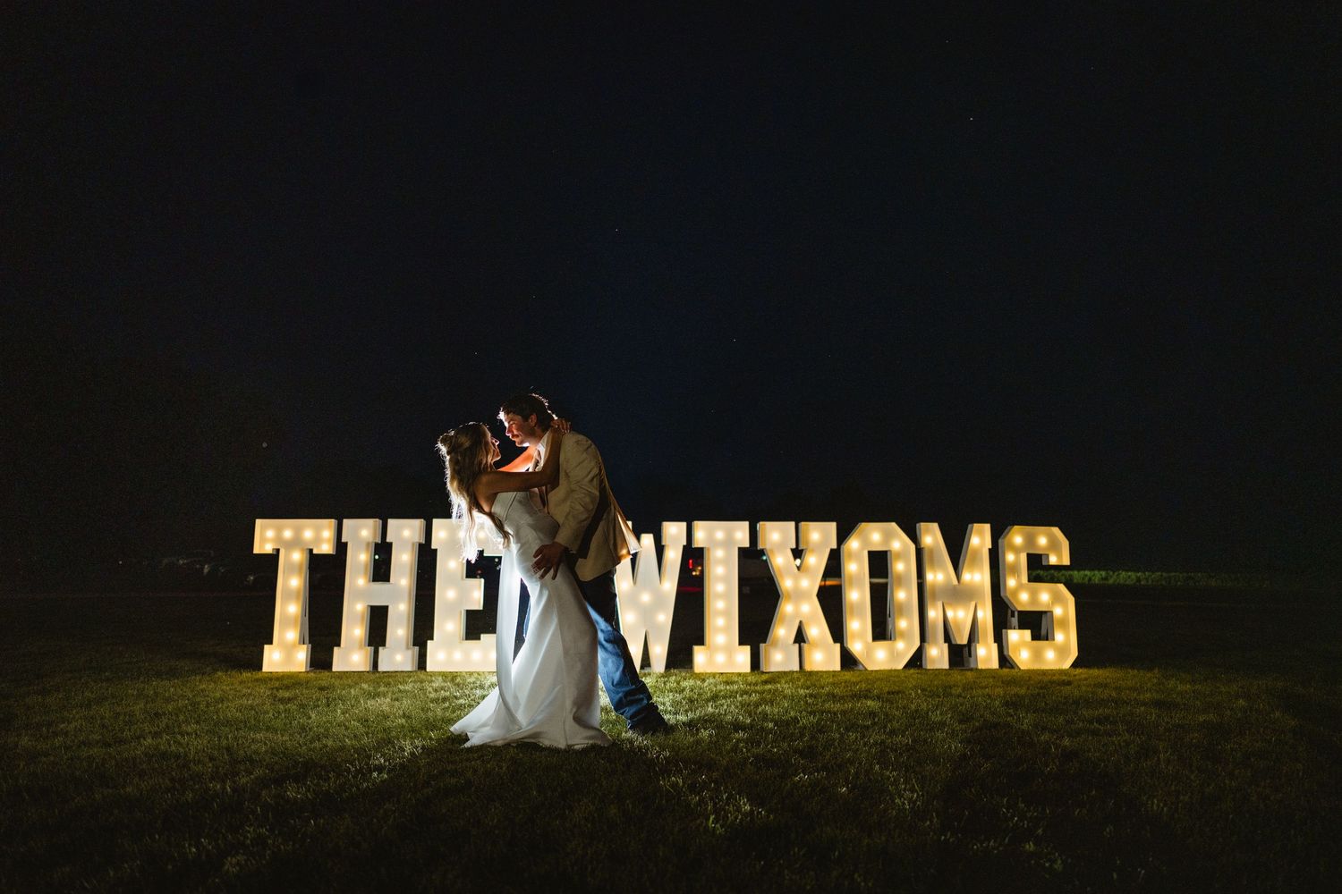 Newlyweds kiss at night in front of illuminated marquee letters spelling THE WIXOMS on a dark outdoor lawn.