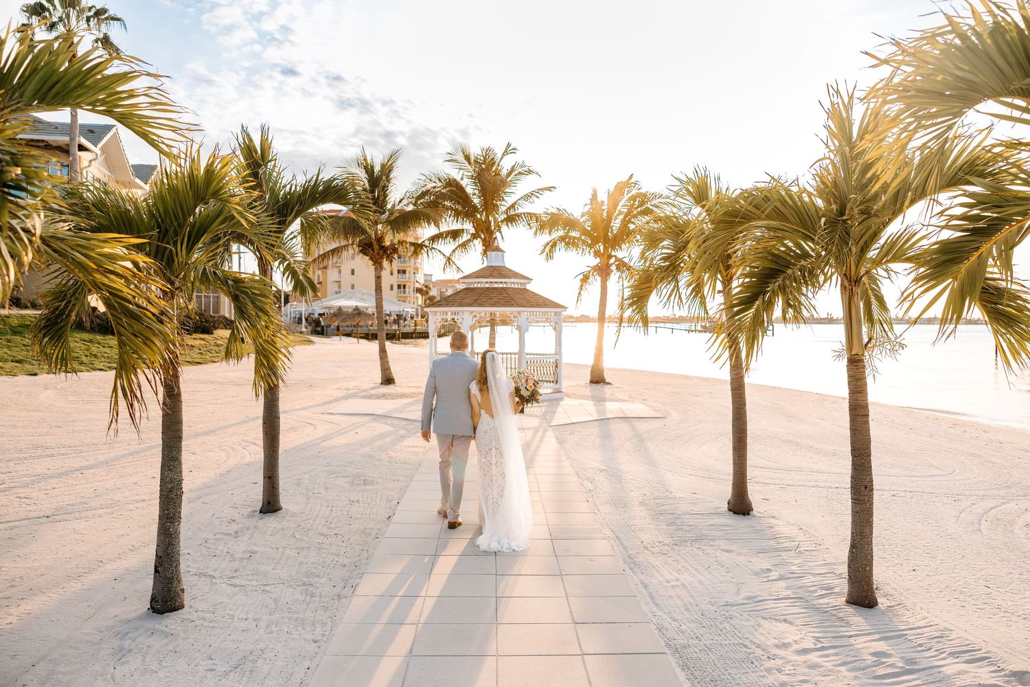 Newlywed couple walking hand-in-hand along a sunny beachside promenade lined with palm trees at golden hour.