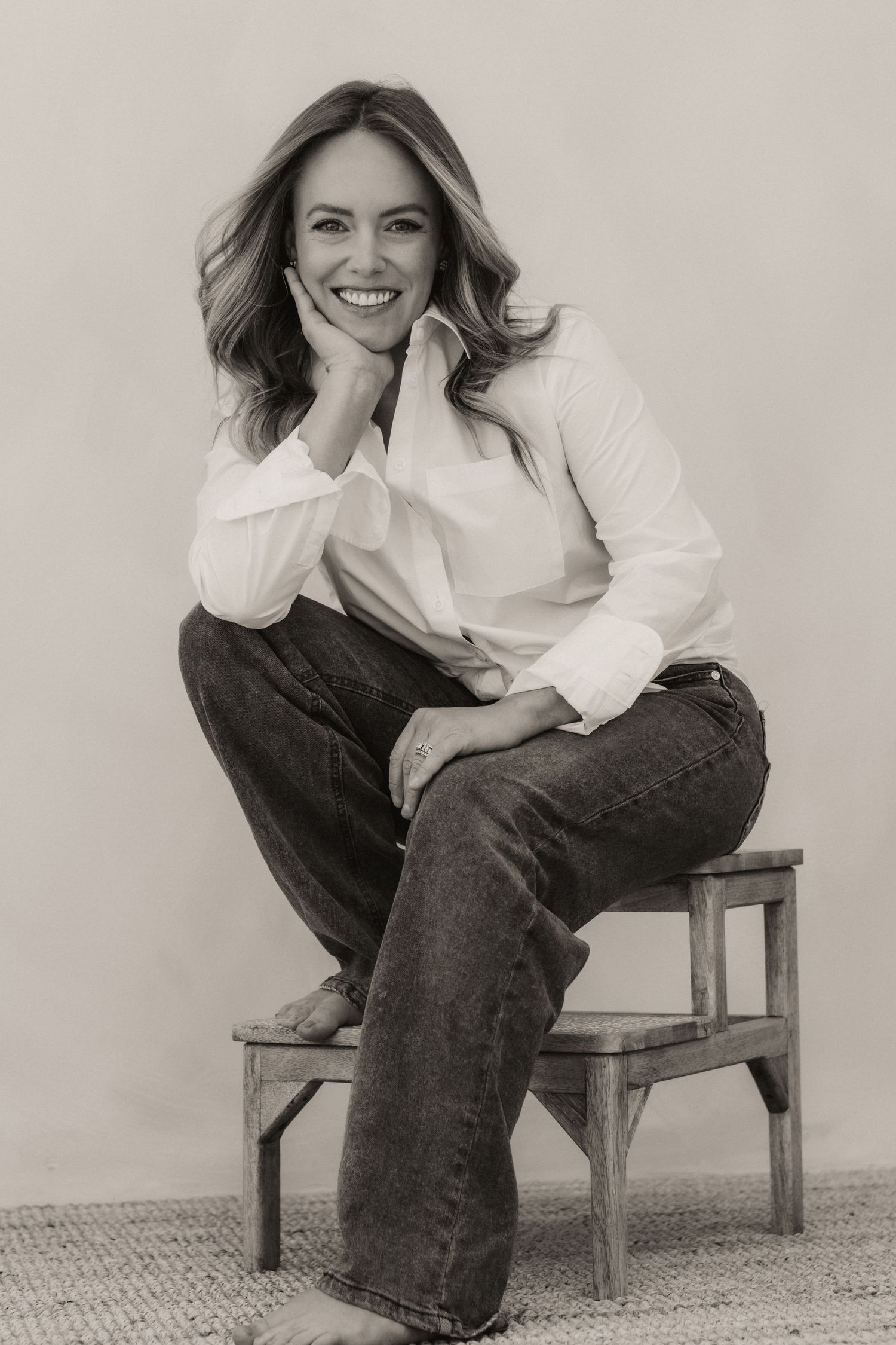 Black and white portrait of a smiling woman in white shirt and jeans sitting on a wooden stool.