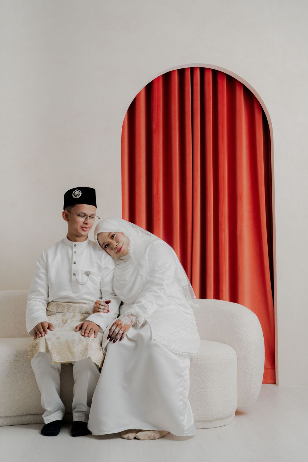 Malaysian couple in traditional white Baju Melayu and hijab wedding attire seated together before a red arched curtain backdrop.