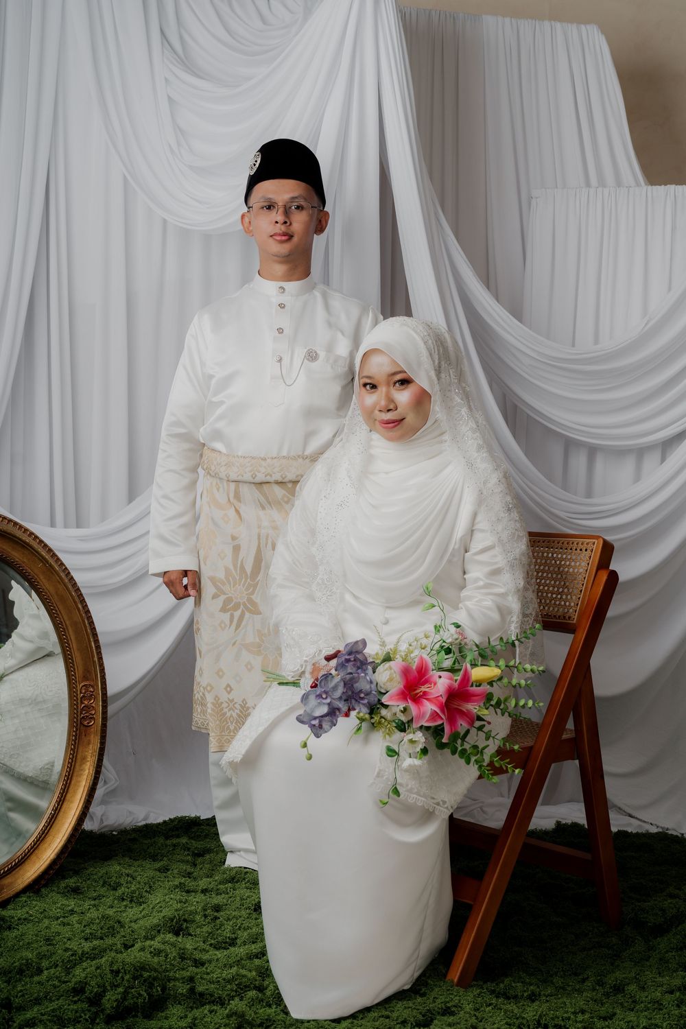 Muslim couple in white traditional wedding attire, bride holding colorful bouquet, posing before draped white fabric backdrop.