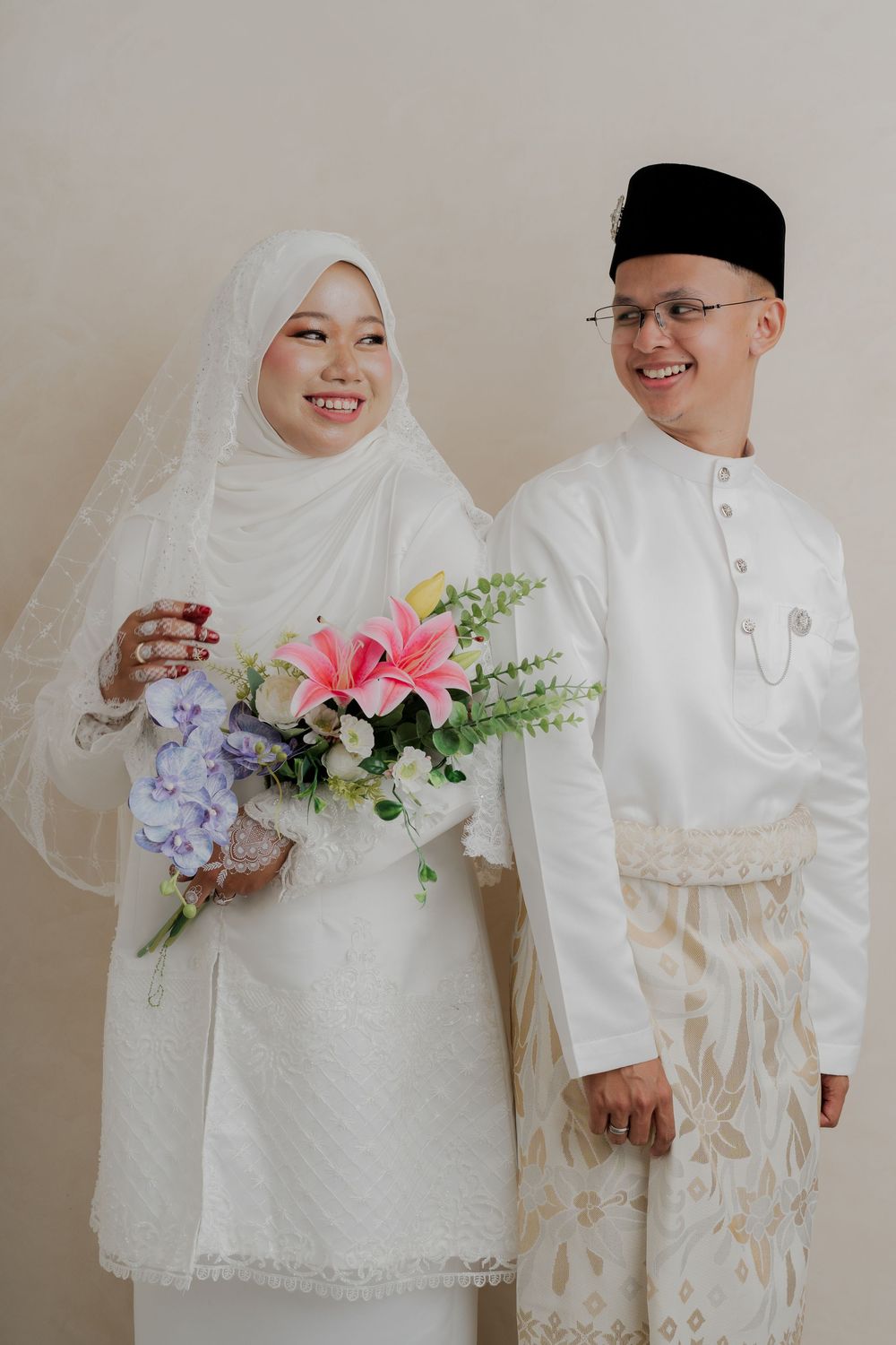 Muslim couple in white traditional wedding attire, bride holding colorful bouquet, groom wearing songkok hat.