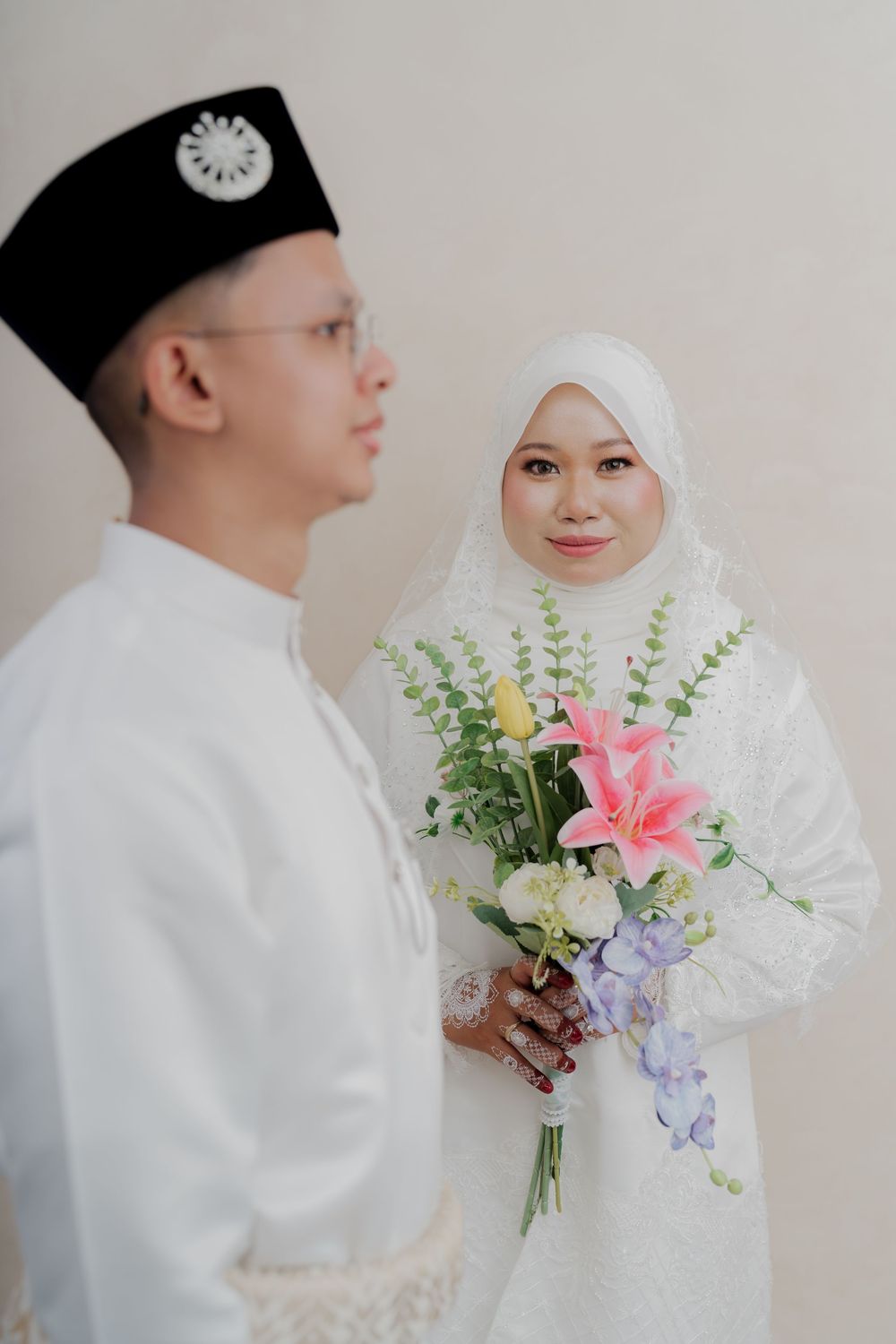 Muslim couple in white wedding attire, bride holding pink lily bouquet, groom wearing songkok hat.