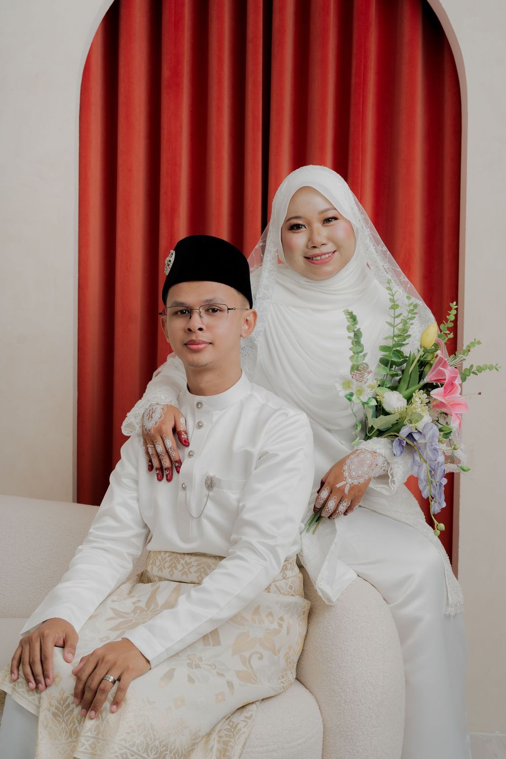Malaysian Muslim couple in white traditional wedding attire, bride holding bouquet, posing before red curtain backdrop.