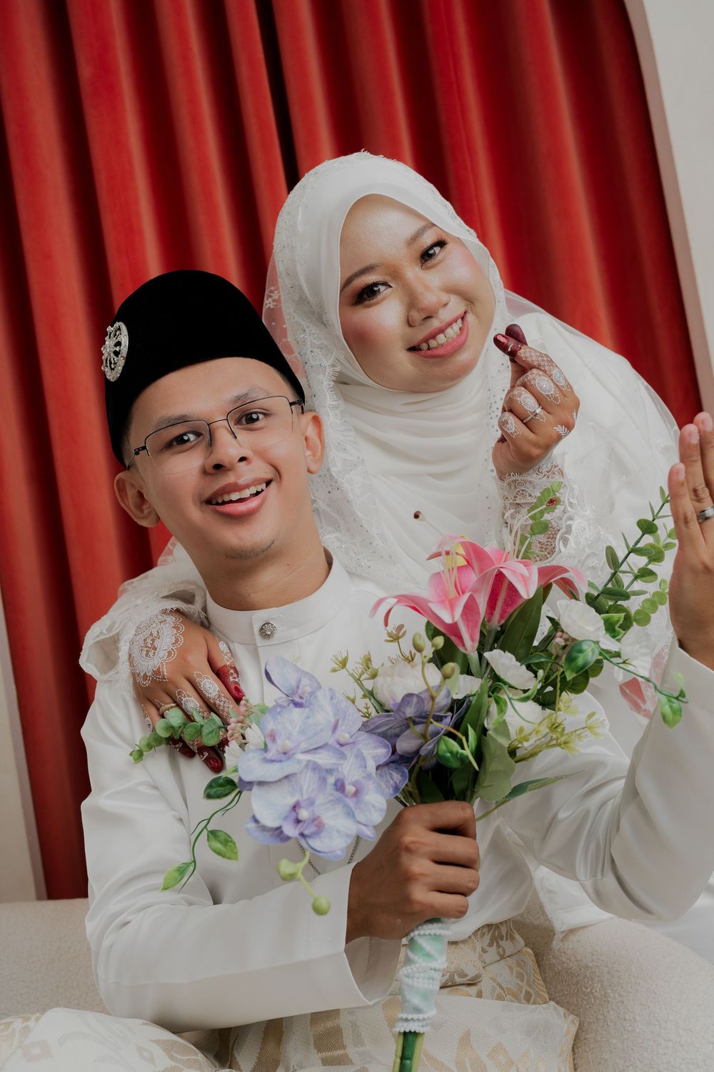A smiling Muslim couple in white traditional attire, groom holding a floral bouquet, bride in hijab, posing together near red curtains.