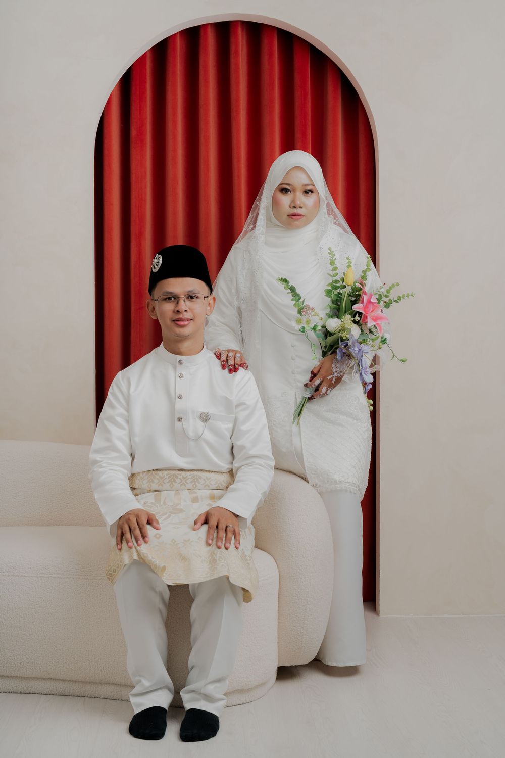 Malay couple in white traditional wedding attire, groom seated wearing songkok, bride standing holding floral bouquet.