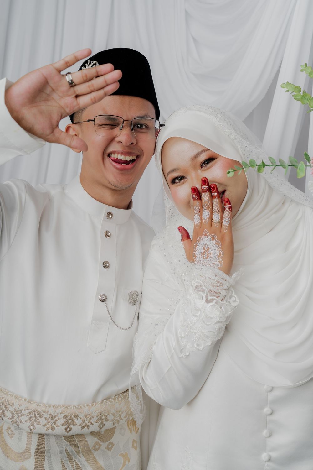 Joyful Malaysian newlyweds in white traditional wedding attire posing playfully, bride showing henna-decorated hands.