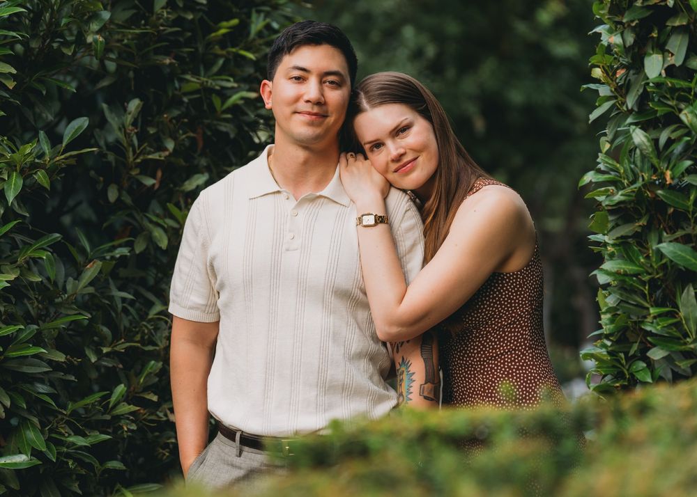 A young couple poses together smiling in front of lush green hedges in an outdoor portrait at inn at little washington