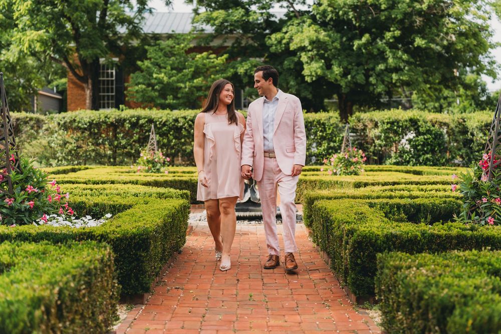 Couple walking hand-in-hand through a formal garden with brick path and hedges, dressed in pink outfits at inn at little washington
