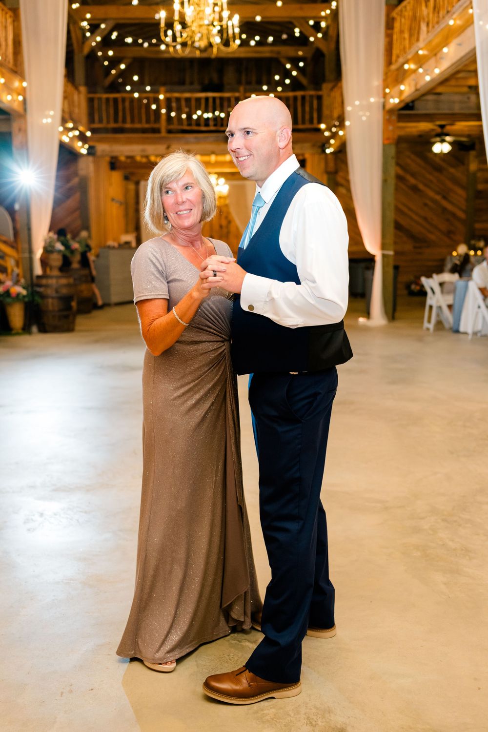 Groom and mother share a tender first dance in a rustic barn wedding venue adorned with warm string lights.