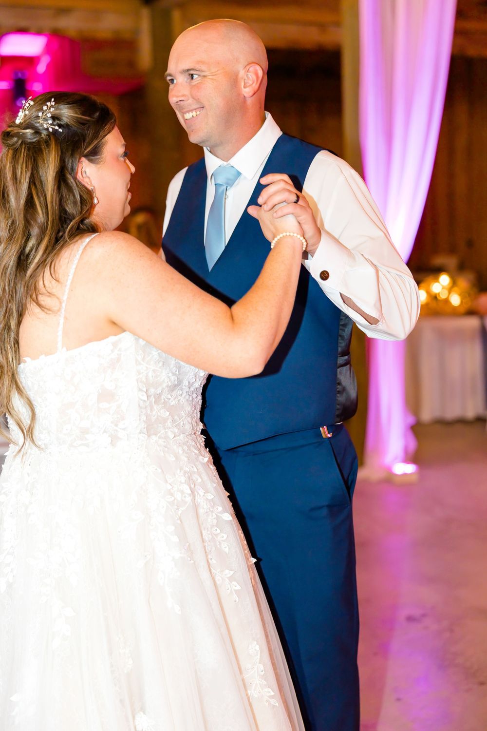 Bride and groom share their first dance at wedding reception, groom in navy suit, bride in white lace gown.