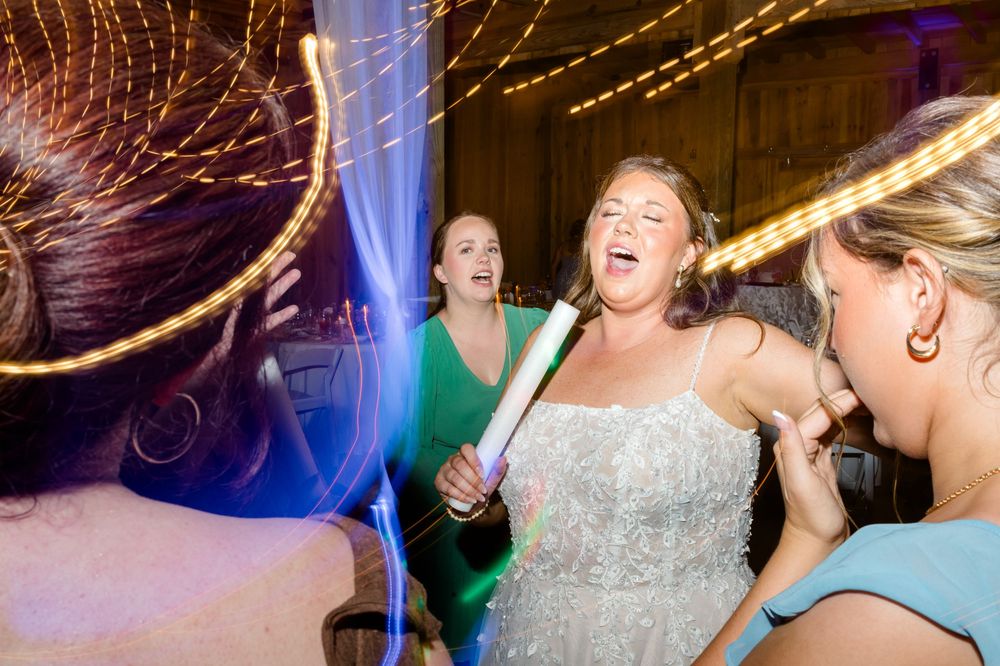 Bride singing with microphone surrounded by guests on wedding dance floor with colorful lights.