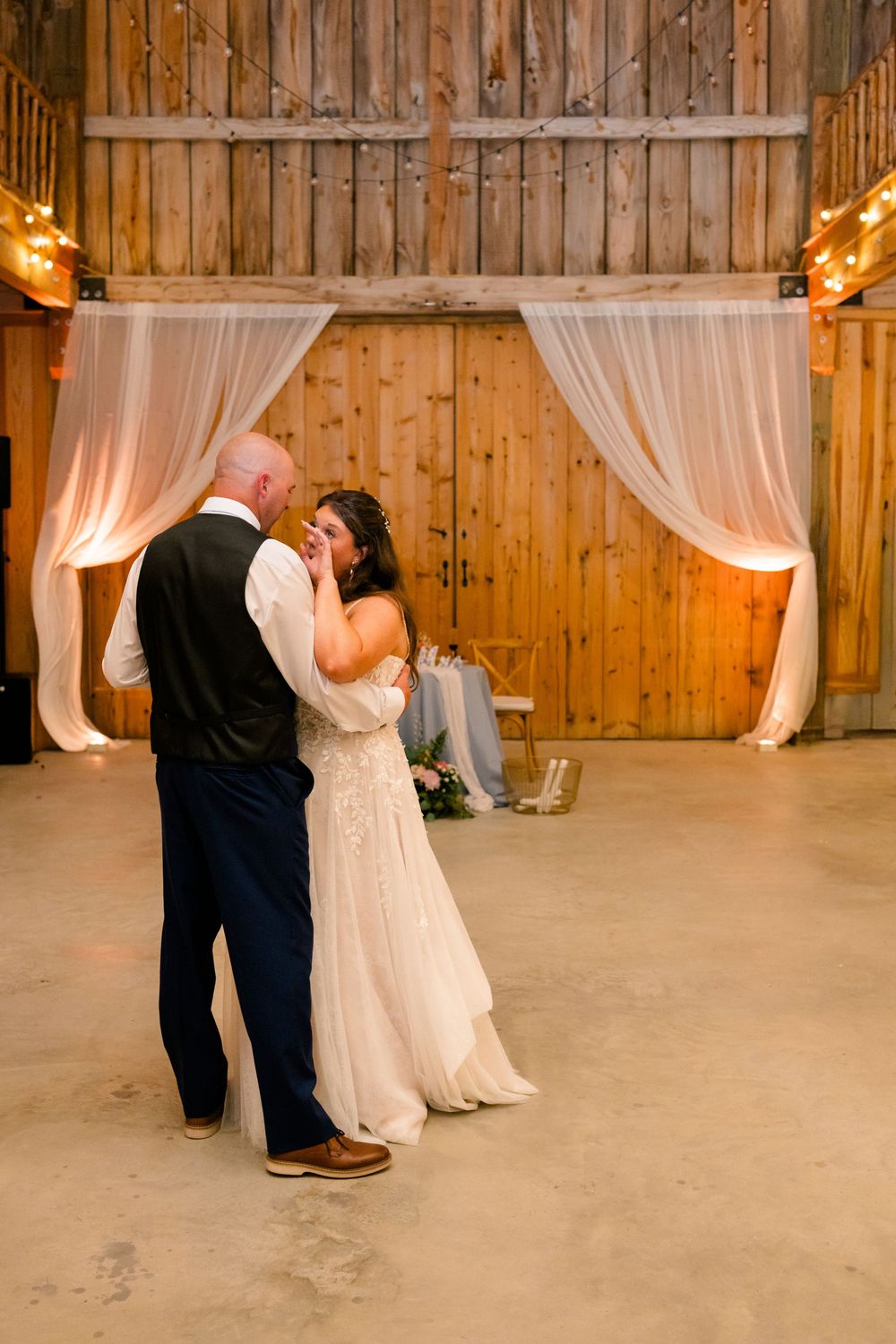 Bride and groom share emotional first dance in rustic barn venue with white draping and warm string lights.