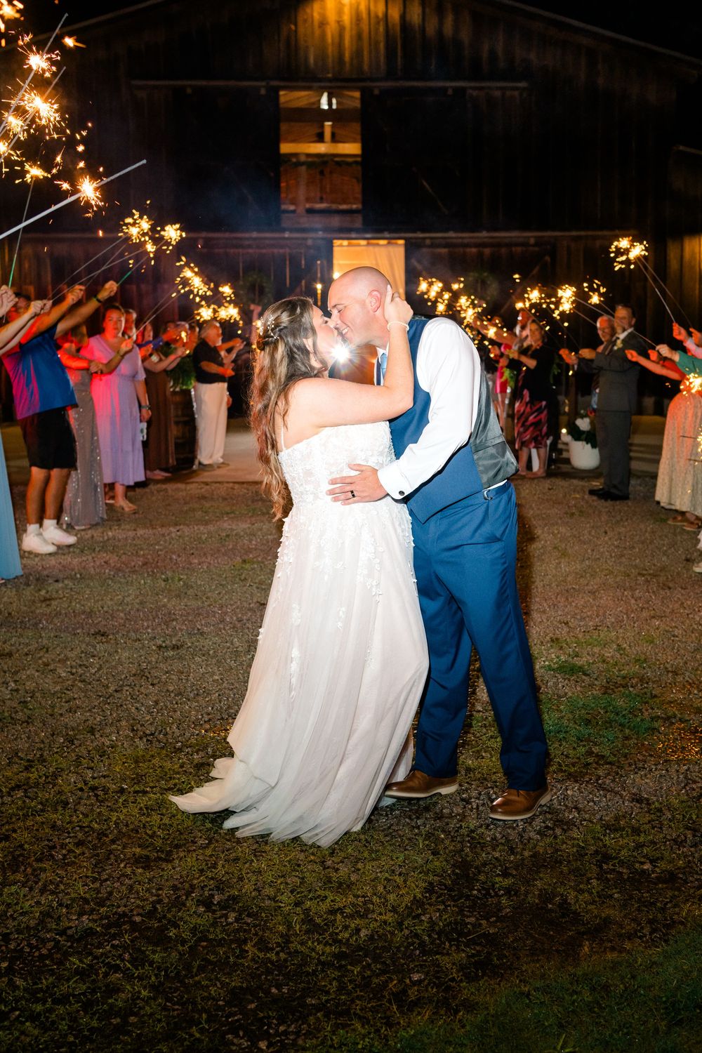 Newlywed couple sharing a kiss during a sparkler send-off exit at their rustic barn wedding reception at night.