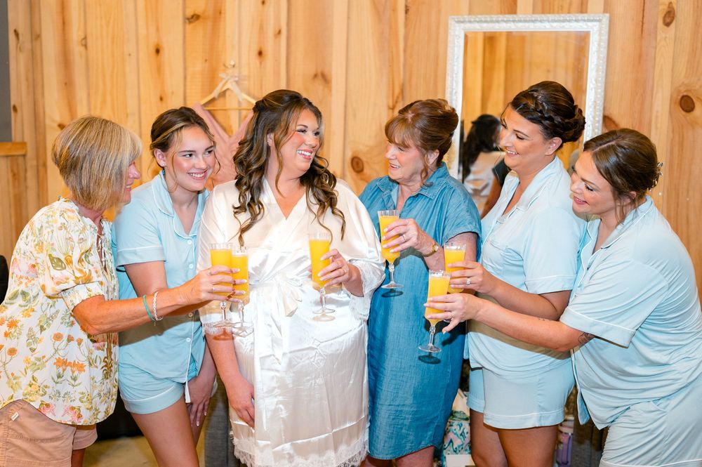 Bride in white robe toasting mimosas with bridesmaids in light blue robes during wedding morning preparations.