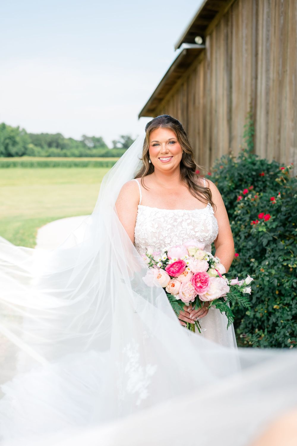 Bride in lace gown with flowing veil holds pink bouquet beside rustic barn and red roses.