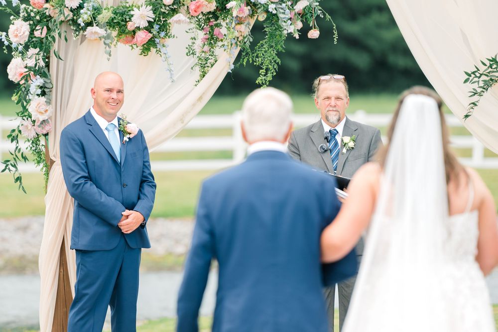 Groom in blue suit smiles as bride walks down the aisle at outdoor wedding ceremony with floral arch.