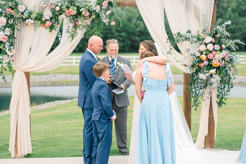 Outdoor wedding ceremony under floral arch with couple, officiant, and child in blue attire on green lawn.