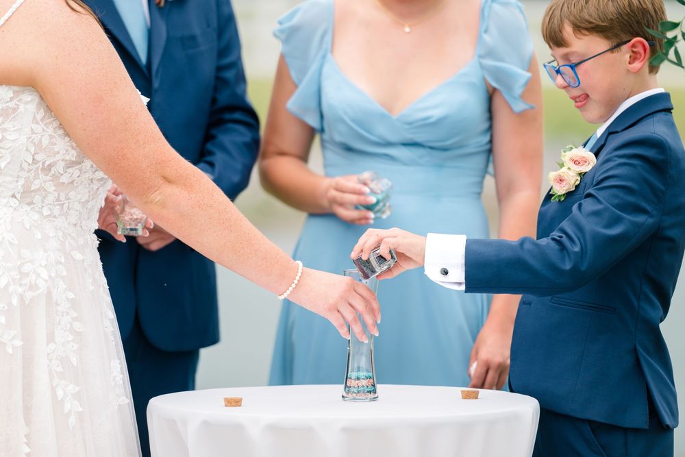 A young boy in a navy suit pours sand during a unity ceremony at a wedding with bridal party members watching.