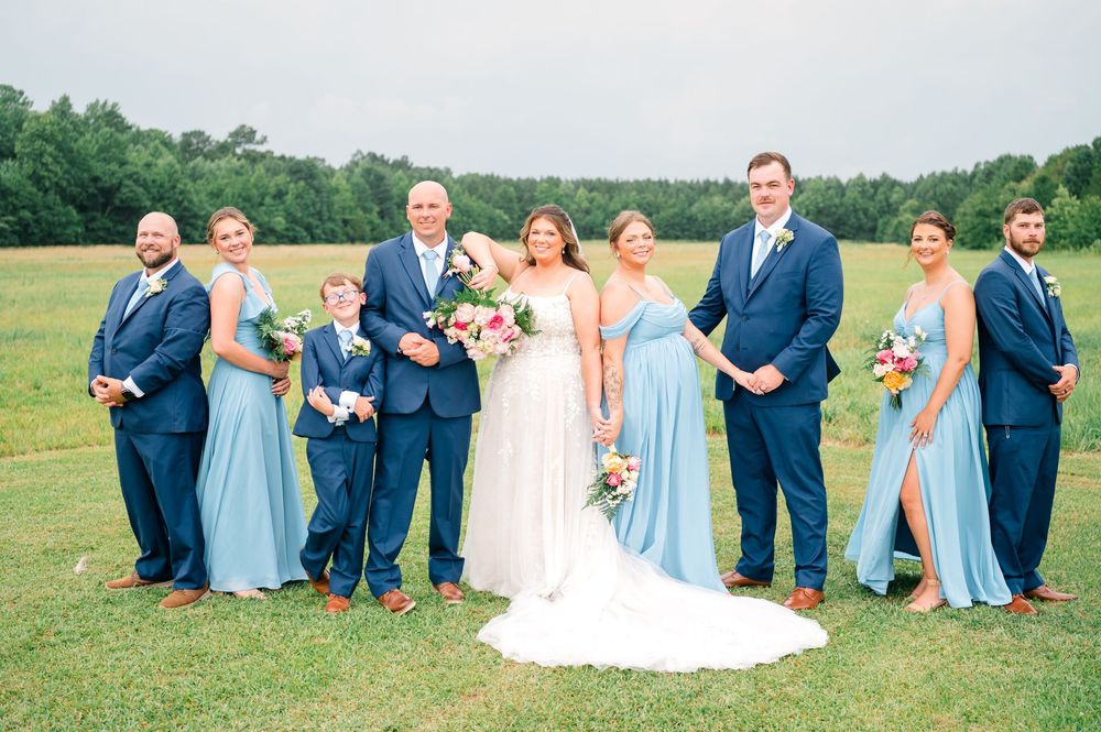 Wedding party posing outdoors in a green field, bridesmaids in blue gowns, groomsmen in navy suits, bride in white dress.