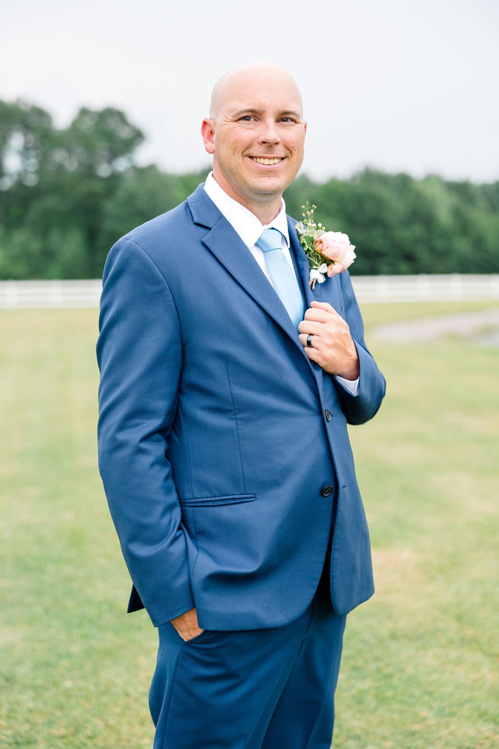 Smiling groom in navy blue suit with boutonniere poses outdoors on green lawn at wedding venue.
