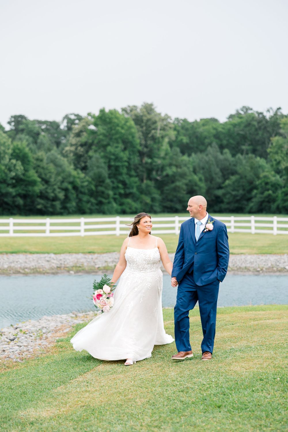 Bride and groom walk hand-in-hand by a pond, she in white gown with bouquet, he in navy suit.