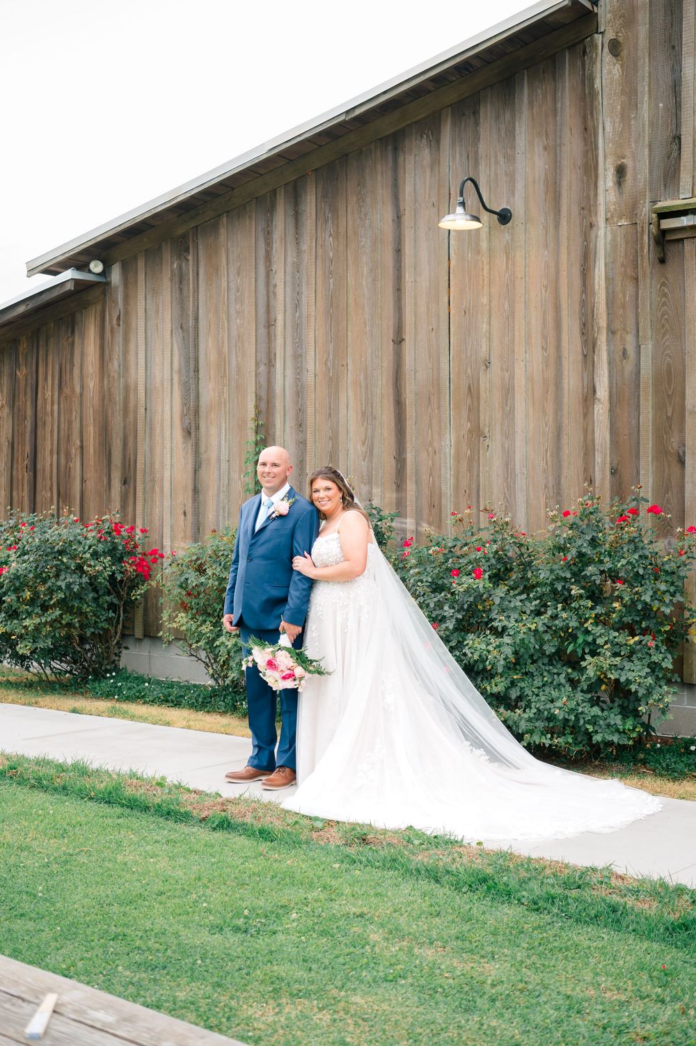 Bride and groom posing together in front of a rustic wooden barn with red roses and green lawn on their wedding day.