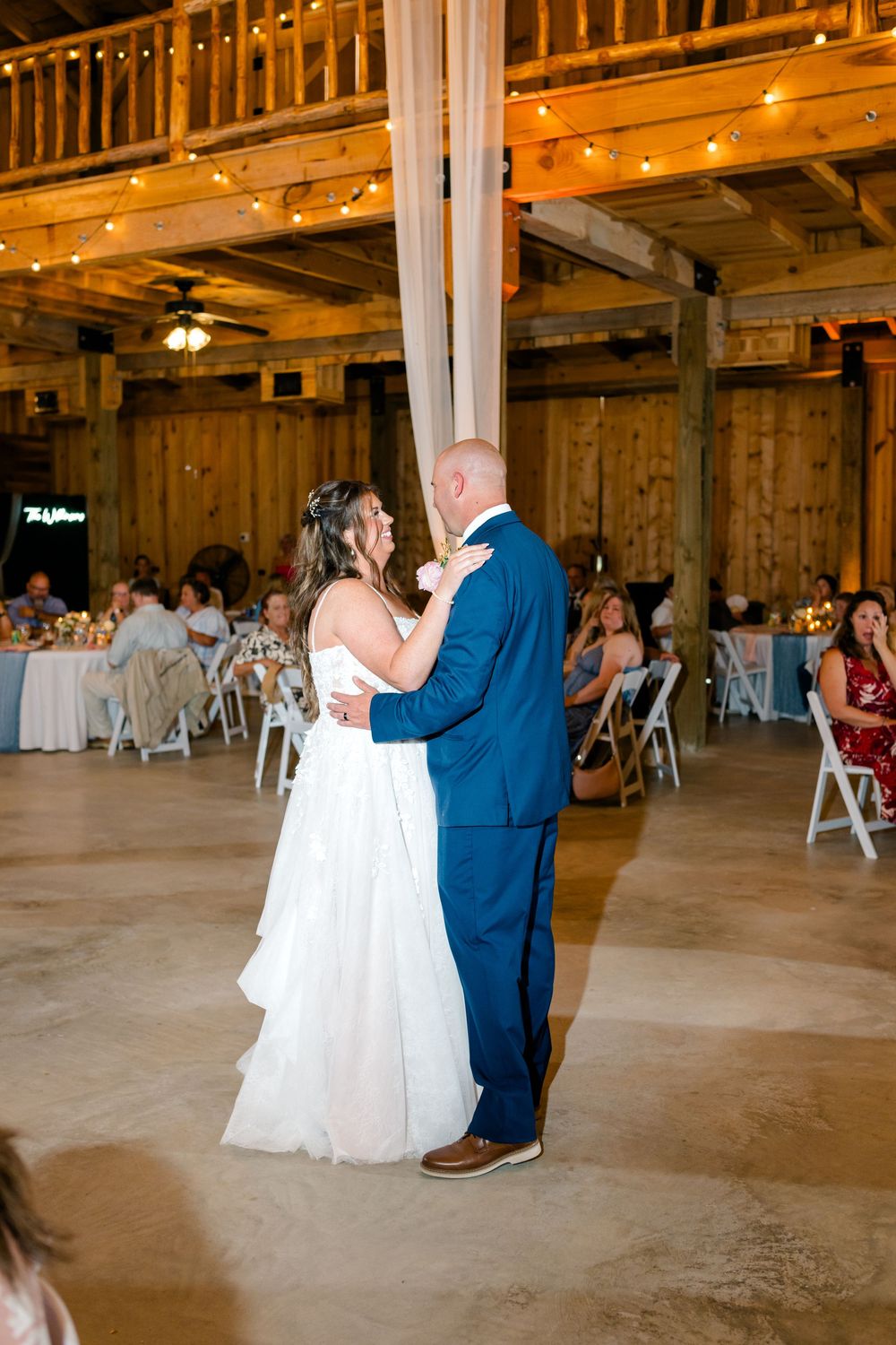 Bride and groom share their first dance in a rustic barn wedding venue with string lights and guests seated nearby.