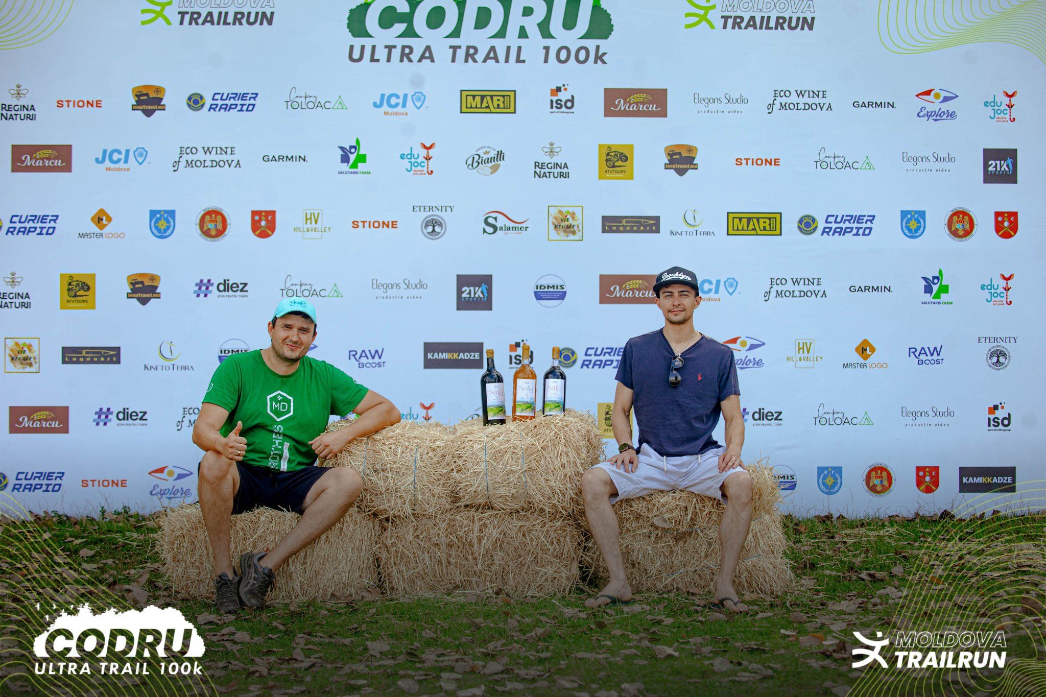 Two male participants pose with wine bottles on hay bales at Codru Ultra Trail 100k race event.