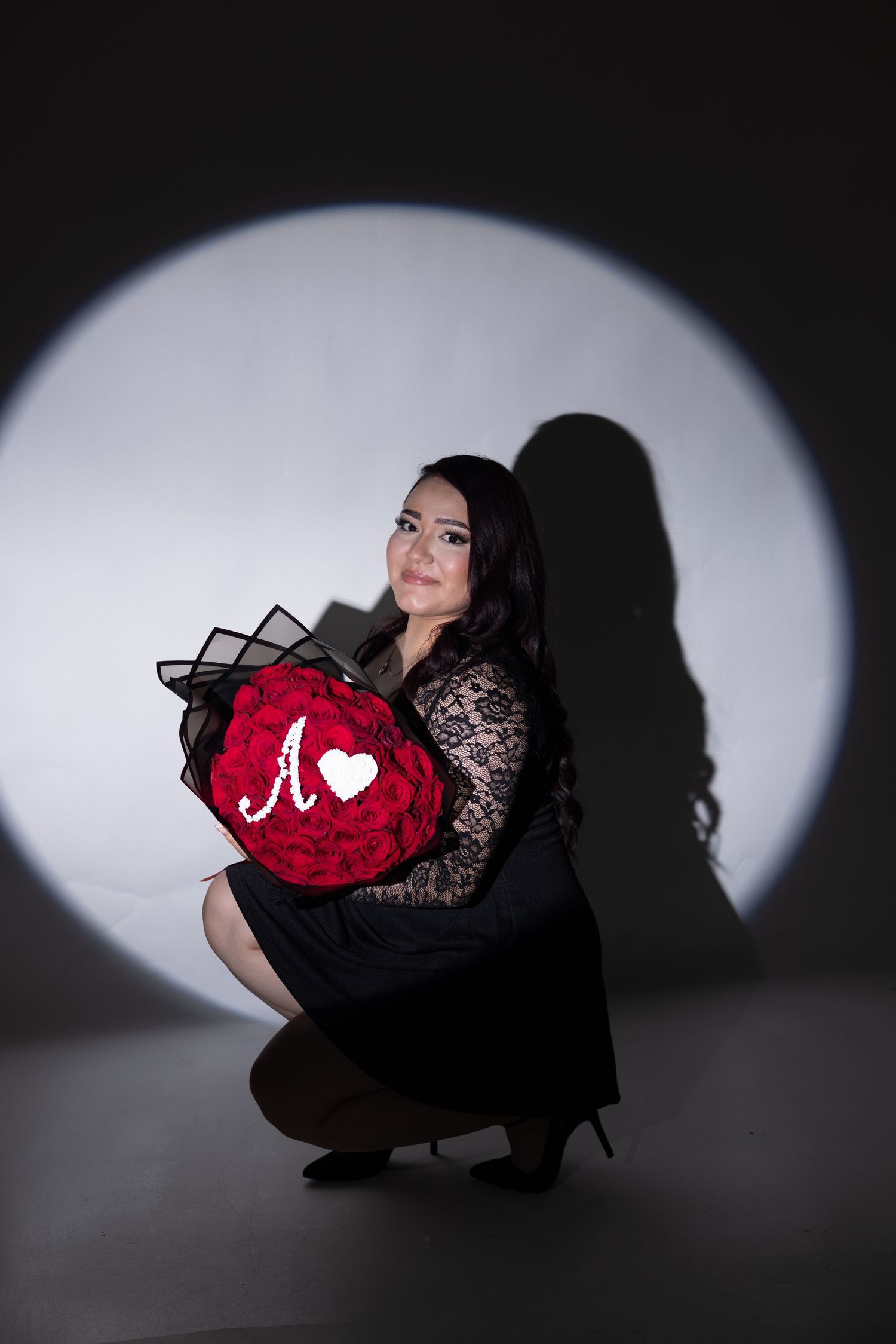 Woman in black lace dress crouching with red rose heart bouquet monogrammed with letter A in dramatic spotlight.