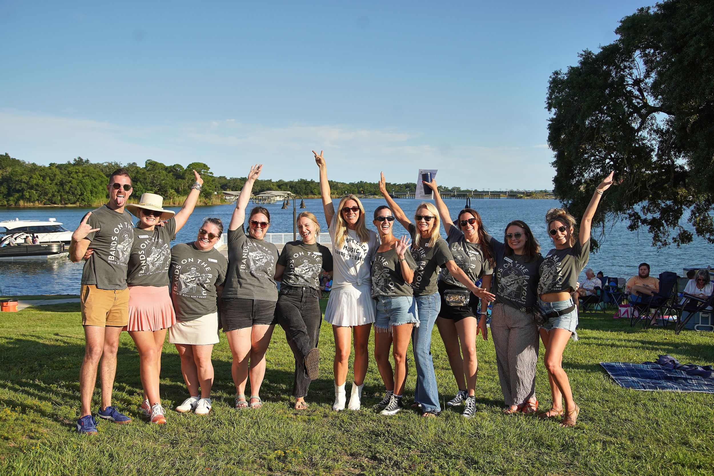 Group of friends in matching gray t-shirts posing and celebrating outdoors near a lake on a sunny day.