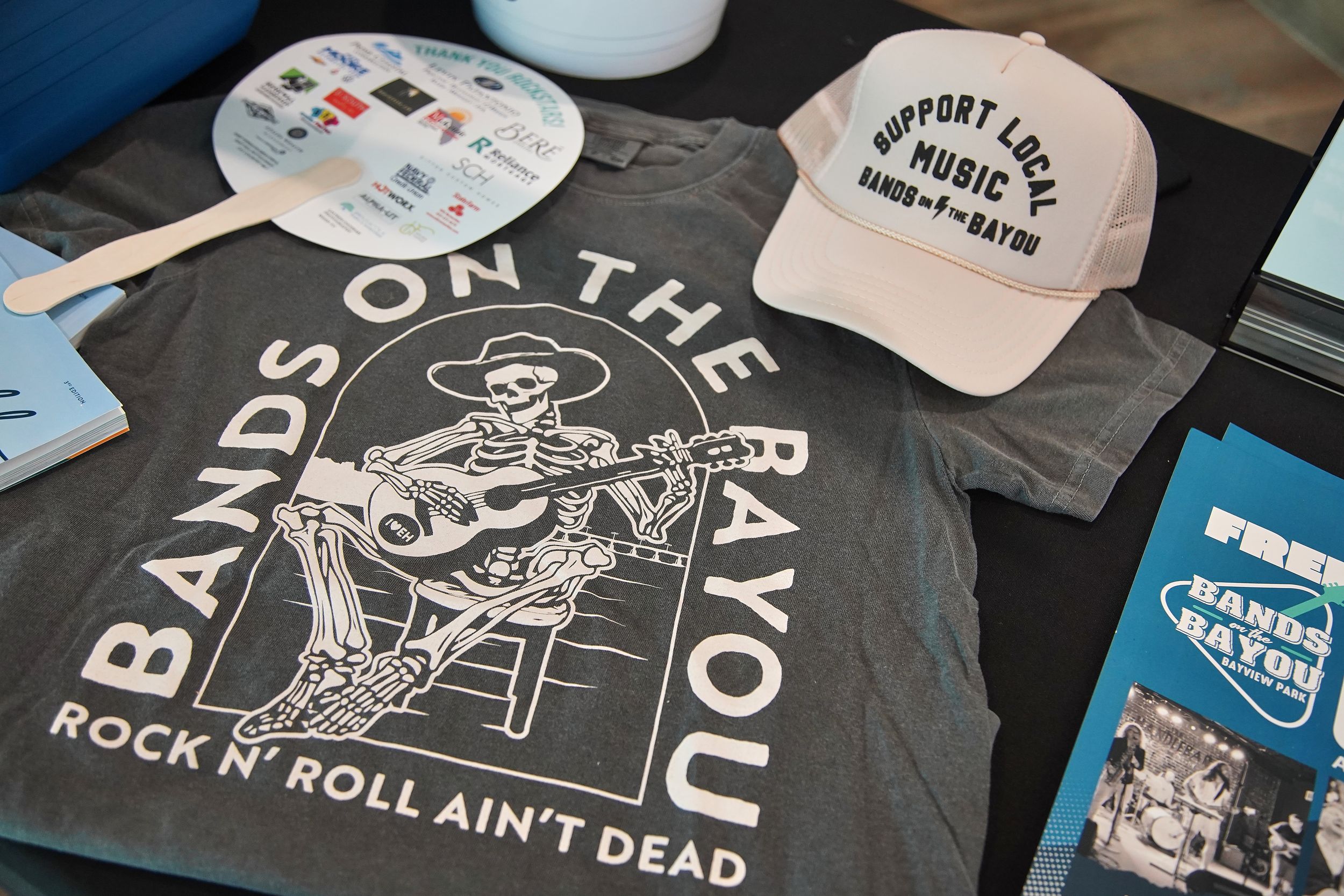 Bands on the Bayou merchandise including a skeleton guitarist t-shirt, trucker hat, and event brochures on display.