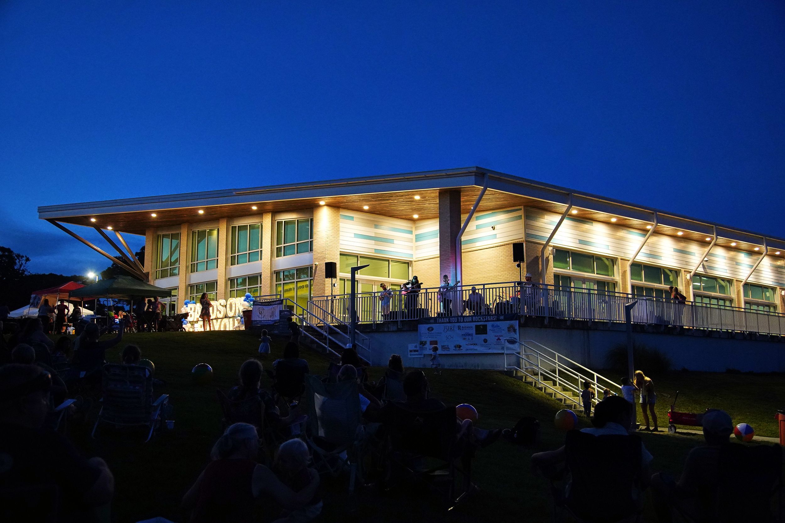 Illuminated modern community center building at dusk with crowd gathered on lawn for outdoor evening event.